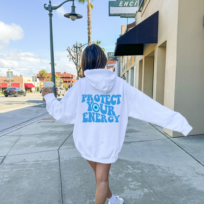 K&ouml;nnte beinhalten: Wei&szlig;er Kapuzenpullover mit einem blauen Augen-Motiv und dem Text "PROTECT YOUR ENERGY" auf der R&uuml;ckseite.