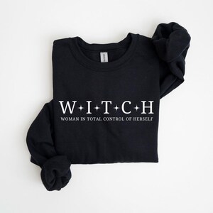 May include: Black long-sleeve sweatshirt with the word "WITCH" in white, with plus signs between each letter. Below the word is the phrase "WOMAN IN TOTAL CONTROL OF HERSELF" in white. The sweatshirt is folded.