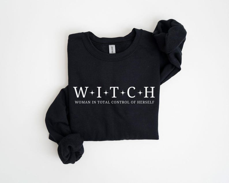 May include: Black long-sleeve sweatshirt with the word "WITCH" in white, with plus signs between each letter. Below the word is the phrase "WOMAN IN TOTAL CONTROL OF HERSELF" in white. The sweatshirt is folded.