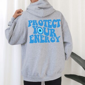 K&ouml;nnte beinhalten: Grauer Kapuzenpullover mit dem Text "PROTECT YOUR ENERGY" in Blau mit einem blauen b&ouml;sen Auge.