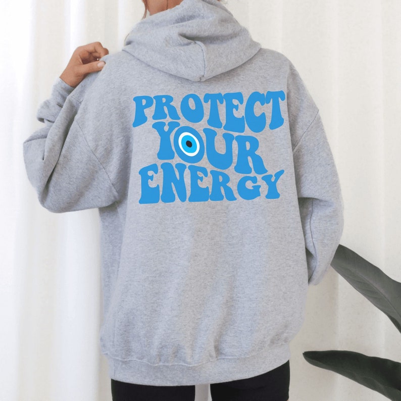 K&ouml;nnte beinhalten: Grauer Kapuzenpullover mit dem Text "PROTECT YOUR ENERGY" in Blau mit einem blauen b&ouml;sen Auge.