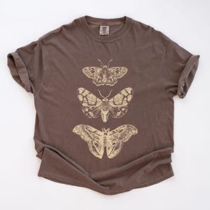 May include: A brown t-shirt featuring three detailed moth illustrations in a light tan color. The moths are arranged vertically, showcasing their intricate wing patterns. The shirt has a classic crew neck and short sleeves.