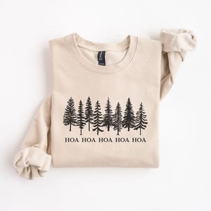 Peut inclure: Un sweat-shirt beige avec un motif noir repr&eacute;sentant une rang&eacute;e de pins. En dessous des arbres, le texte "HOA HOA HOA HOA HOA" est imprim&eacute; en noir. Le sweat-shirt a des manches longues et un col rond.