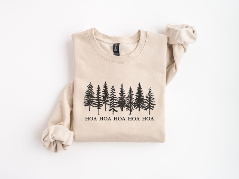 Peut inclure: Un sweat-shirt beige avec un motif noir repr&eacute;sentant une rang&eacute;e de pins. En dessous des arbres, le texte "HOA HOA HOA HOA HOA" est imprim&eacute; en noir. Le sweat-shirt a des manches longues et un col rond.