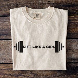 K&ouml;nnte beinhalten: Ein wei&szlig;es T-Shirt mit dem Text "LIFT LIKE A GIRL" in Schwarz gedruckt, mit einer Hantelgrafik auf jeder Seite des Textes.