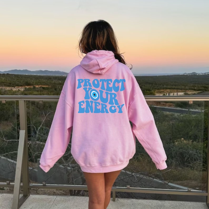 K&ouml;nnte beinhalten: Rosa Kapuzenpullover mit dem Text "PROTECT YOUR ENERGY" in Blau mit einem blauen b&ouml;sen Auge.