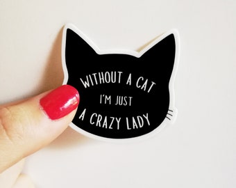 Puede incluir: Una pegatina negra con forma de gato con texto blanco que dice "WITHOUT A CAT I'M JUST A CRAZY LADY". La pegatina está sujeta por un dedo con esmalte de uñas rojo.