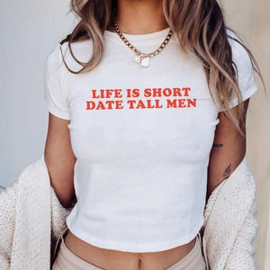 Könnte beinhalten: Weißes Crop-Top mit dem roten Text "LIFE IS SHORT DATE TALL MEN". Das Shirt hat kurze Ärmel und einen Rundhalsausschnitt. Eine goldene Halskette wird um den Hals getragen.