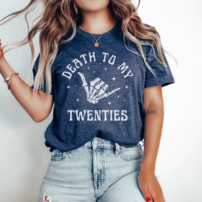 May include: A navy blue t-shirt with a white graphic of a skeleton hand making a shaka sign. The text "Death to my twenties" is printed in white above the hand.