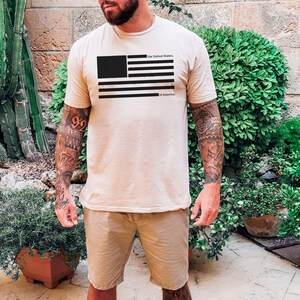 Peut inclure: T-shirt beige avec un drapeau am&eacute;ricain noir et blanc et le texte "the United States, of America". La personne porte un short kaki. L'image est prise &agrave; l'ext&eacute;rieur, avec des plantes en arri&egrave;re-plan.