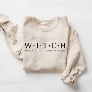 May include: A beige sweatshirt with the word "WITCH" spelled out in black capital letters, with the phrase "WOMAN IN TOTAL CONTROL OF HERSELF" underneath. The sleeves are rolled up, and the sweatshirt is laid flat on a white surface.