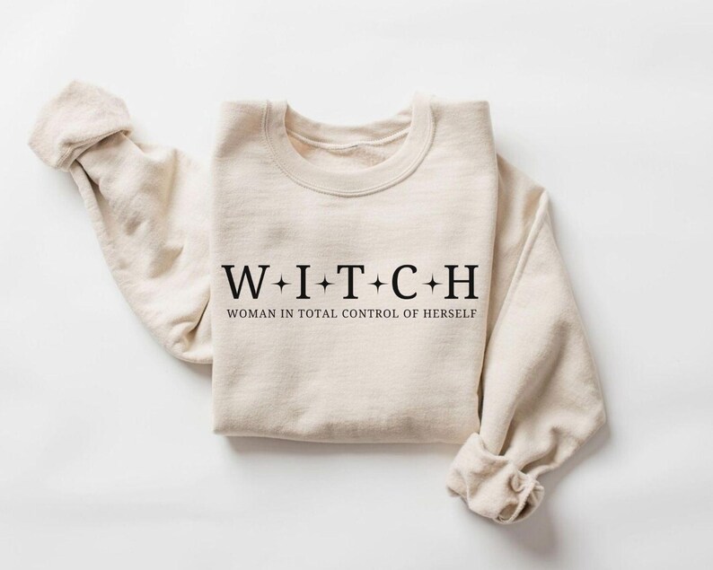 May include: A beige sweatshirt with the word "WITCH" spelled out in black capital letters, with the phrase "WOMAN IN TOTAL CONTROL OF HERSELF" underneath. The sleeves are rolled up, and the sweatshirt is laid flat on a white surface.
