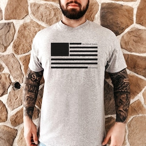 Peut inclure: T-shirt gris clair avec un motif noir du drapeau am&eacute;ricain et les mots "the United States" et "of America". Le t-shirt a un col rond et des manches courtes.