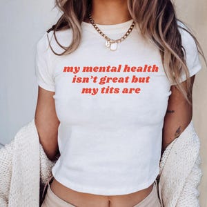 Könnte beinhalten: Cremefarbenes Crop-Top mit dem roten Text "my mental health isn't great but my tits are". Das Shirt wird mit beigefarbener Hose und einer goldenen Halskette kombiniert.