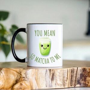 May include: A white ceramic mug with a black handle and rim featuring a green matcha drink illustration with a smiling face and the text YOU MEAN SO MATCHA TO ME printed in green letters on the front of the mug surface.