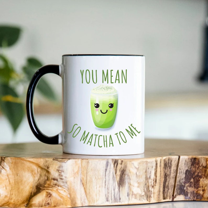 May include: A white ceramic mug with a black handle and rim featuring a green matcha drink illustration with a smiling face and the text YOU MEAN SO MATCHA TO ME printed in green letters on the front of the mug surface.
