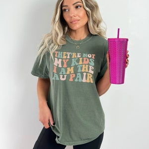 Pode incluir: Uma camiseta verde com o texto "They're not my kids I am the au pair" em uma fonte retr&ocirc; com cores rosa, laranja e azul.