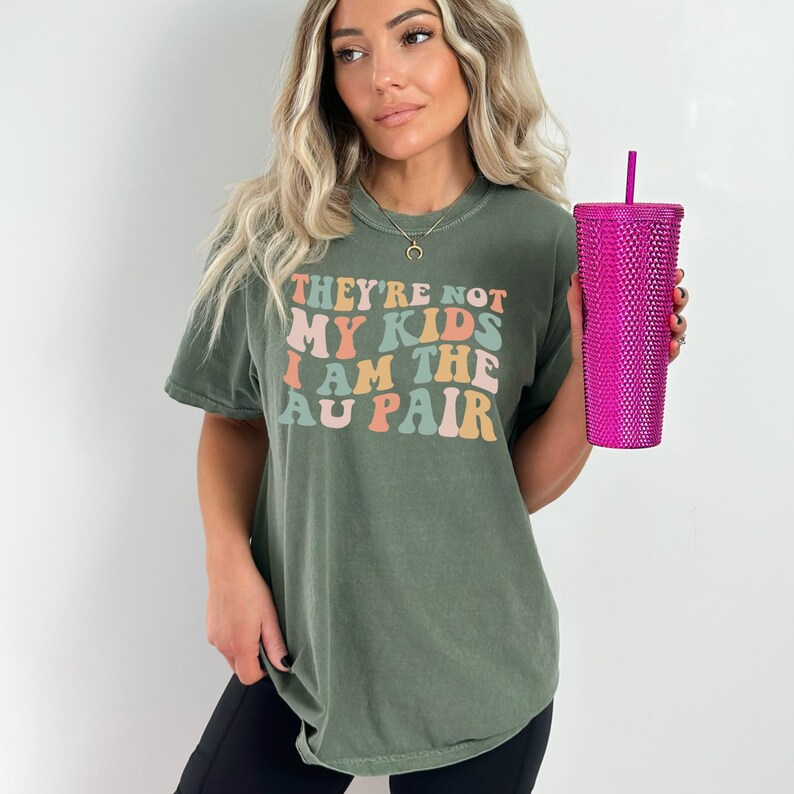 Pode incluir: Uma camiseta verde com o texto "They're not my kids I am the au pair" em uma fonte retr&ocirc; com cores rosa, laranja e azul.
