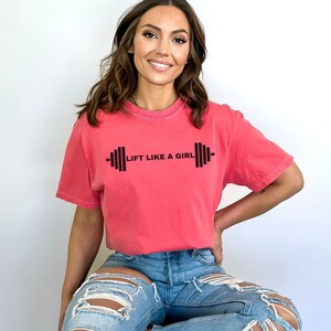 K&ouml;nnte beinhalten: Eine Frau tr&auml;gt ein pinkfarbenes T-Shirt mit dem Text "Lift Like A Girl" und einer schwarzen Hantelgrafik. Sie tr&auml;gt eine blaue Jeans mit Destroyed-Details.