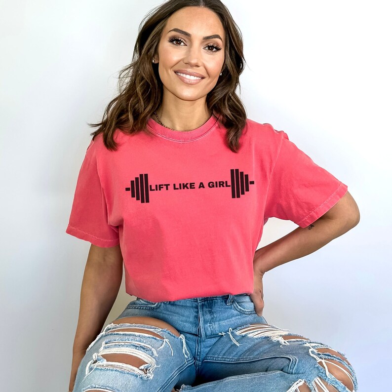K&ouml;nnte beinhalten: Eine Frau tr&auml;gt ein pinkfarbenes T-Shirt mit dem Text "Lift Like A Girl" und einer schwarzen Hantelgrafik. Sie tr&auml;gt eine blaue Jeans mit Destroyed-Details.