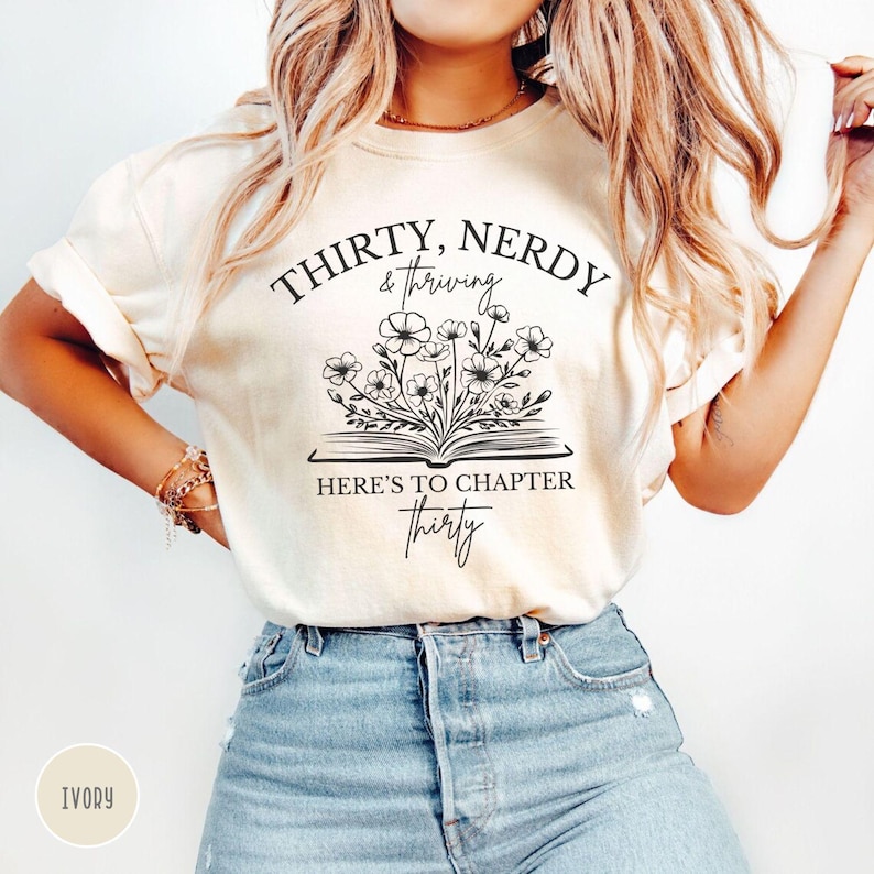 May include: Ivory t-shirt with black text that reads "Thirty, Nerdy & Thriving. Here's to Chapter Thirty."  The text is surrounded by a black outline of a book with flowers growing out of it.