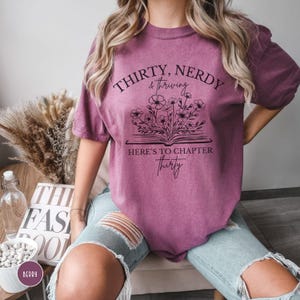 May include: A mauve t-shirt with a graphic design of a book with flowers growing out of it. The text on the shirt reads "Thirty, Nerdy & Thriving. Here's to Chapter Thirty."