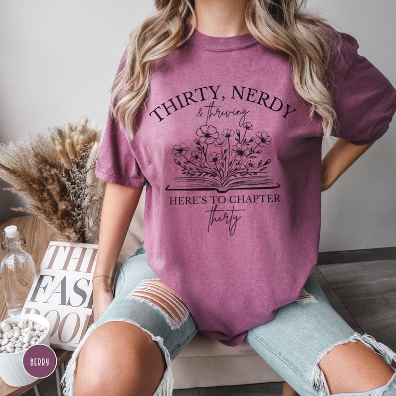May include: A mauve t-shirt with a graphic design of a book with flowers growing out of it. The text on the shirt reads "Thirty, Nerdy & Thriving. Here's to Chapter Thirty."