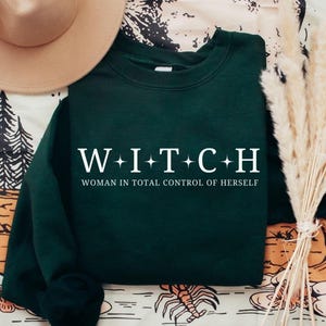 May include: Dark green sweatshirt with the word "WITCH" spelled out in white capital letters, with a plus sign between each letter. Below the word is the phrase "WOMAN IN TOTAL CONTROL OF HERSELF" in white.