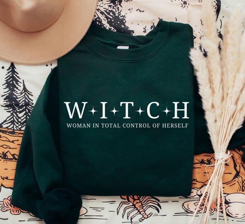 May include: Dark green sweatshirt with the word "WITCH" spelled out in white capital letters, with a plus sign between each letter. Below the word is the phrase "WOMAN IN TOTAL CONTROL OF HERSELF" in white.