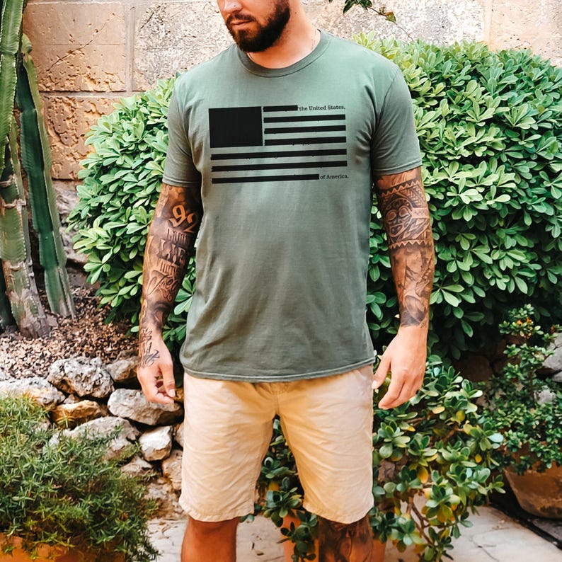 Peut inclure: T-shirt vert olive avec un drapeau am&eacute;ricain noir imprim&eacute;. Le texte "the United States, of America" est imprim&eacute; au-dessus et en dessous du drapeau. Le t-shirt est port&eacute; avec un short kaki.