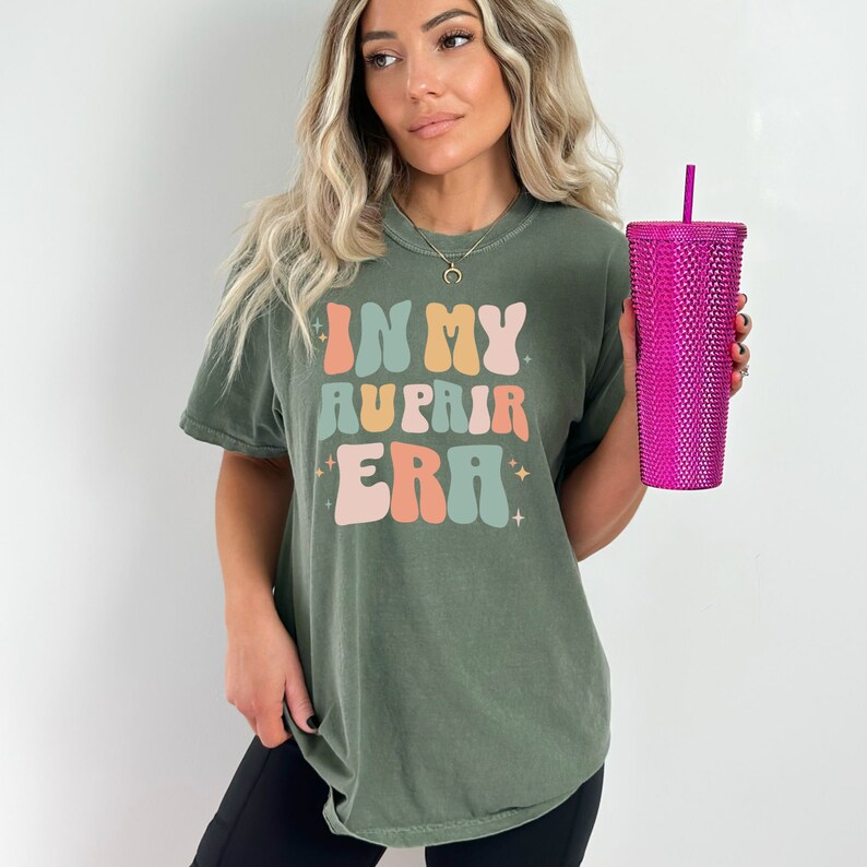 May include: A green t-shirt with the text "In My Au Pair Era" in colourful retro font. The shirt is being worn by a person holding a pink studded tumbler.