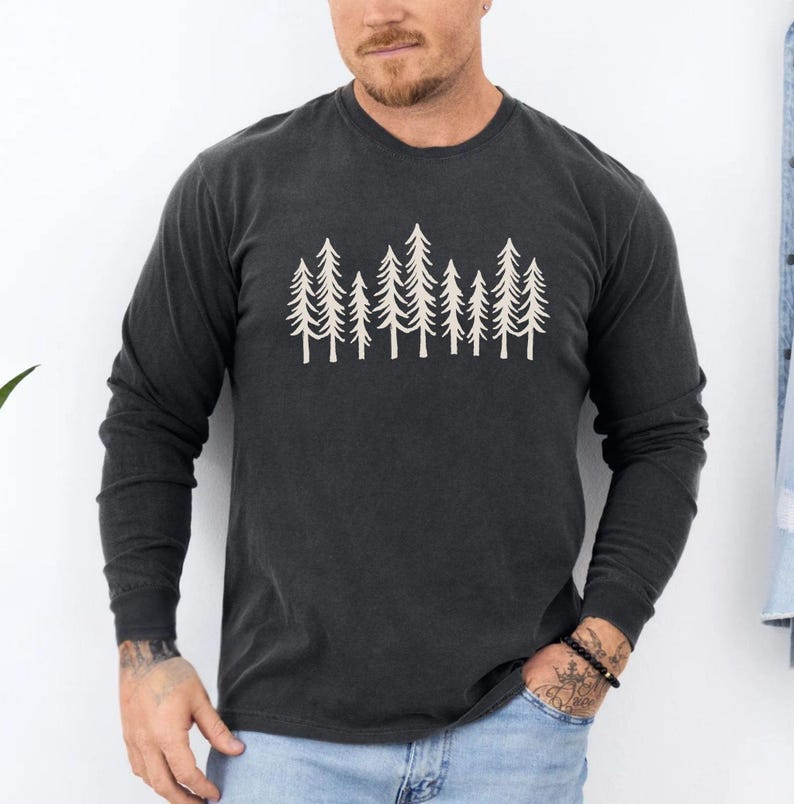 K&ouml;nnte beinhalten: Dunkelgraues Langarmshirt mit cremefarbenem Baumlinien-Grafik. Das Shirt hat einen Rundhalsausschnitt und eine lockere Passform. Das Grafikdesign ist eine einfache, minimalistische Illustration von mehreren Kiefern.