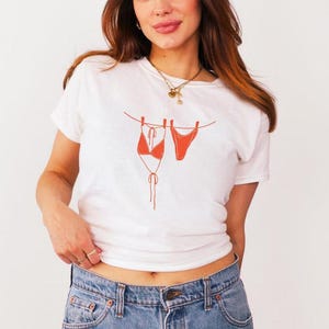 Könnte beinhalten: Weißes T-Shirt mit einem orangefarbenen Bikini-Motiv, das an einer Wäscheleine hängt. Das Shirt wird mit einer blauen Jeans kombiniert. Das Design ist eine einfache Strichzeichnung, die eine minimalistische Ästhetik schafft. Das Thema ist Sommer- und Strandmode.