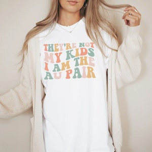 Pode incluir: Camiseta branca com o texto "They're not my kids I am the au pair" em fonte retr&ocirc; colorida.