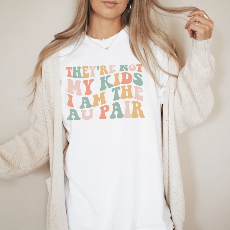 Pode incluir: Camiseta branca com o texto "They're not my kids I am the au pair" em fonte retr&ocirc; colorida.