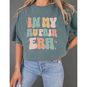 May include: A teal blue t-shirt with the text "In My Au Pair Era" in a retro, colourful font.