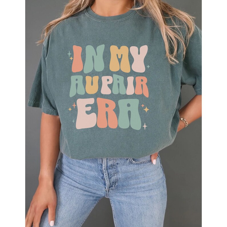 May include: A teal blue t-shirt with the text "In My Au Pair Era" in a retro, colourful font.