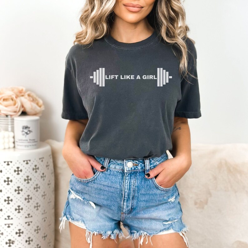 K&ouml;nnte beinhalten: Ein dunkelgraues T-Shirt mit einer wei&szlig;en Grafik einer Langhantel und dem Text "LIFT LIKE A GIRL".