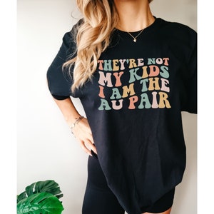 Pode incluir: Camiseta preta com um gr&aacute;fico colorido que diz "They're not my kids I am the au pair".