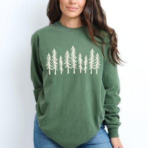 K&ouml;nnte beinhalten: Ein lang&auml;rmeliges, waldgr&uuml;nes Shirt mit einer wei&szlig;en Grafik von stilisierten immergr&uuml;nen B&auml;umen. Das Shirt hat einen Rundhalsausschnitt und eine lockere Passform. Das Model tr&auml;gt blaue Jeans.