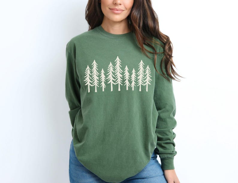 K&ouml;nnte beinhalten: Ein lang&auml;rmeliges, waldgr&uuml;nes Shirt mit einer wei&szlig;en Grafik von stilisierten immergr&uuml;nen B&auml;umen. Das Shirt hat einen Rundhalsausschnitt und eine lockere Passform. Das Model tr&auml;gt blaue Jeans.