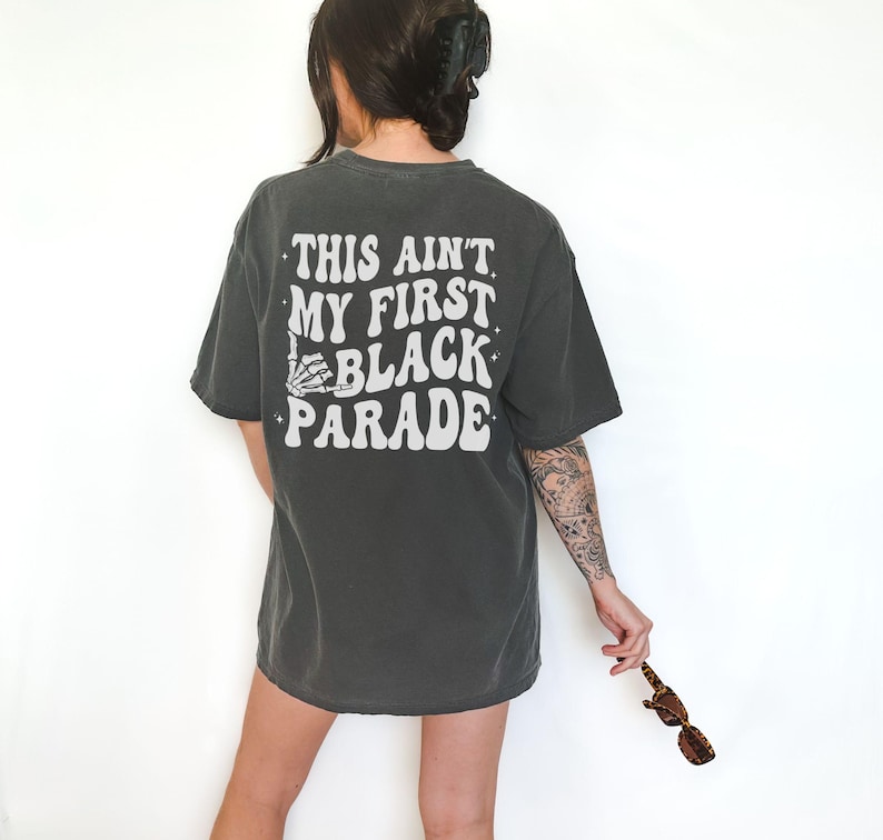 May include: A dark gray t-shirt with the text "This ain't my first black parade" printed in white. The text is in a retro font style.