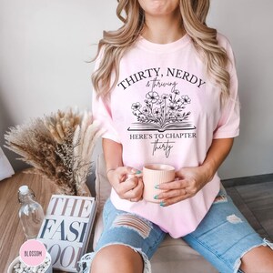 May include: A pink t-shirt with the text "Thirty, Nerdy & Thriving" and "Here's to Chapter Thirty" with a black and white illustration of a book with flowers growing out of it.