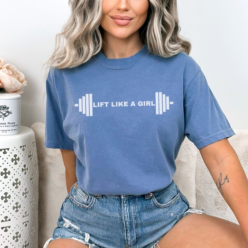 K&ouml;nnte beinhalten: Ein blaues T-Shirt mit dem Text "LIFT LIKE A GIRL" und einer gedruckten Hantelgrafik auf der Vorderseite.