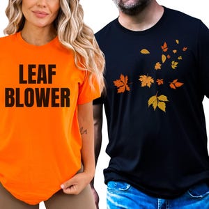 May include: Two short-sleeved t-shirts. One is orange with the text "LEAF BLOWER" in black. The other is black with a design of falling autumn leaves in shades of orange and yellow. Both shirts are casual wear.