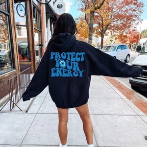 K&ouml;nnte beinhalten: Schwarzer Kapuzenpullover mit einem blauen und wei&szlig;en Augen-Motiv und dem Text "PROTECT YOUR ENERGY" in einer Retro-Schrift.
