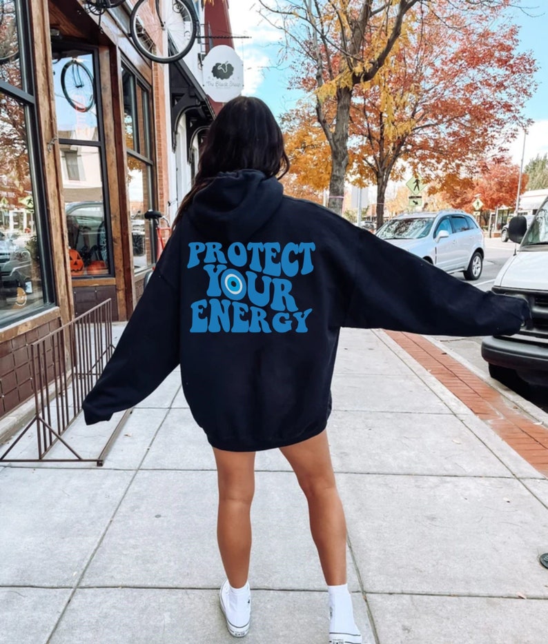 K&ouml;nnte beinhalten: Schwarzer Kapuzenpullover mit einem blauen und wei&szlig;en Augen-Motiv und dem Text "PROTECT YOUR ENERGY" in einer Retro-Schrift.