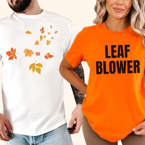 May include: Two short-sleeved t-shirts. One is white with orange and yellow autumn leaves. The other is orange with the words "LEAF BLOWER" in black block letters. The shirts are casual wear.