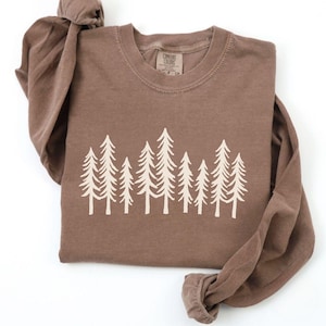 K&ouml;nnte beinhalten: Ein braunes Langarmshirt mit Rundhalsausschnitt. Das Shirt hat eine cremefarbene Grafik von stilisierten Kiefern auf der Brust. Das Kleidungsst&uuml;ck ist sauber auf einer wei&szlig;en Oberfl&auml;che gefaltet.