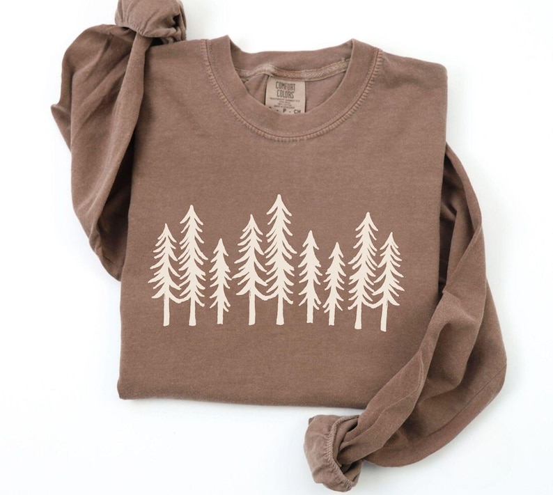 K&ouml;nnte beinhalten: Ein braunes Langarmshirt mit Rundhalsausschnitt. Das Shirt hat eine cremefarbene Grafik von stilisierten Kiefern auf der Brust. Das Kleidungsst&uuml;ck ist sauber auf einer wei&szlig;en Oberfl&auml;che gefaltet.